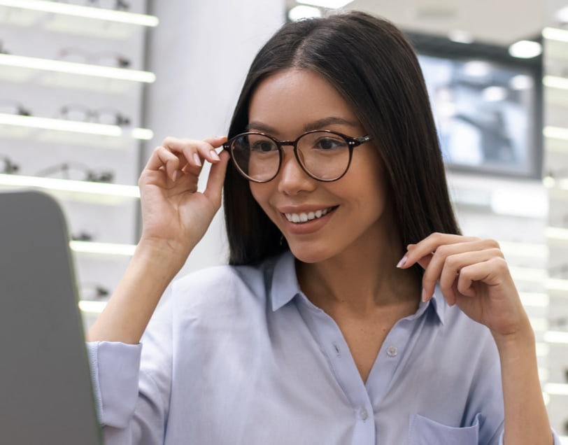 Young woman trying on eyeglasses, looking into a mirror and smiling