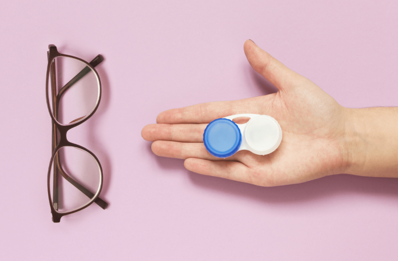A pair of glasses sitting against a pink background, with someone holding their hand out with a contact lens case in the palm of their hand