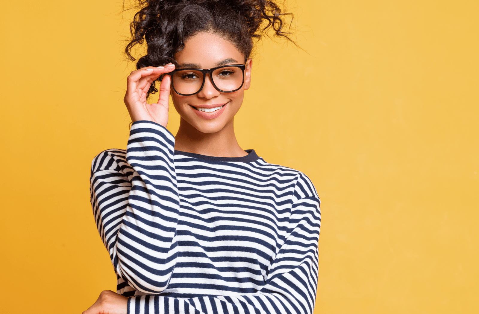 A young woman wearing large, black-rimmed glasses and a black and white striped shirt against a yellow backdrop