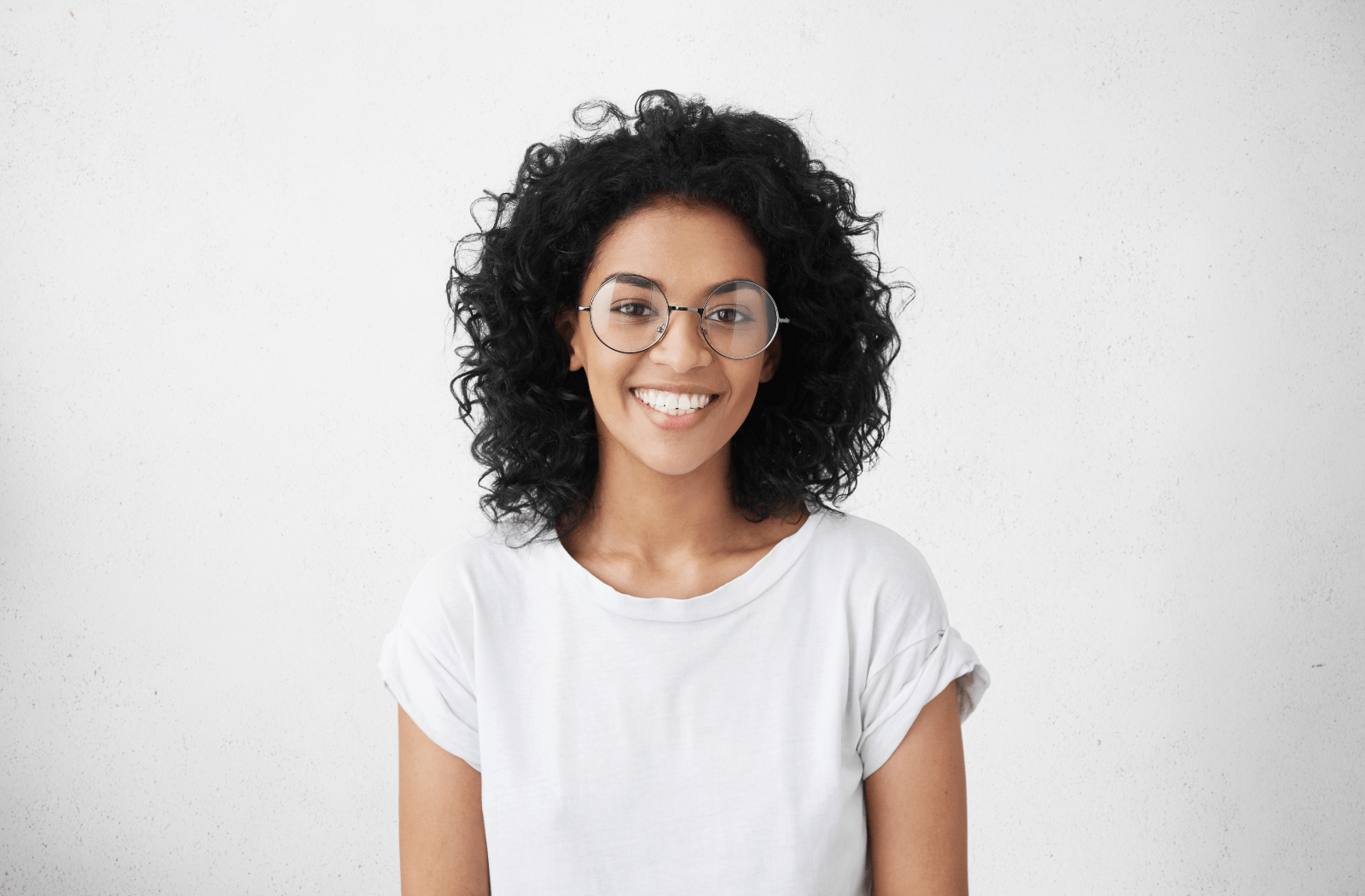 A young woman wearing prescription glasses smiling at the camera and standing against a white background