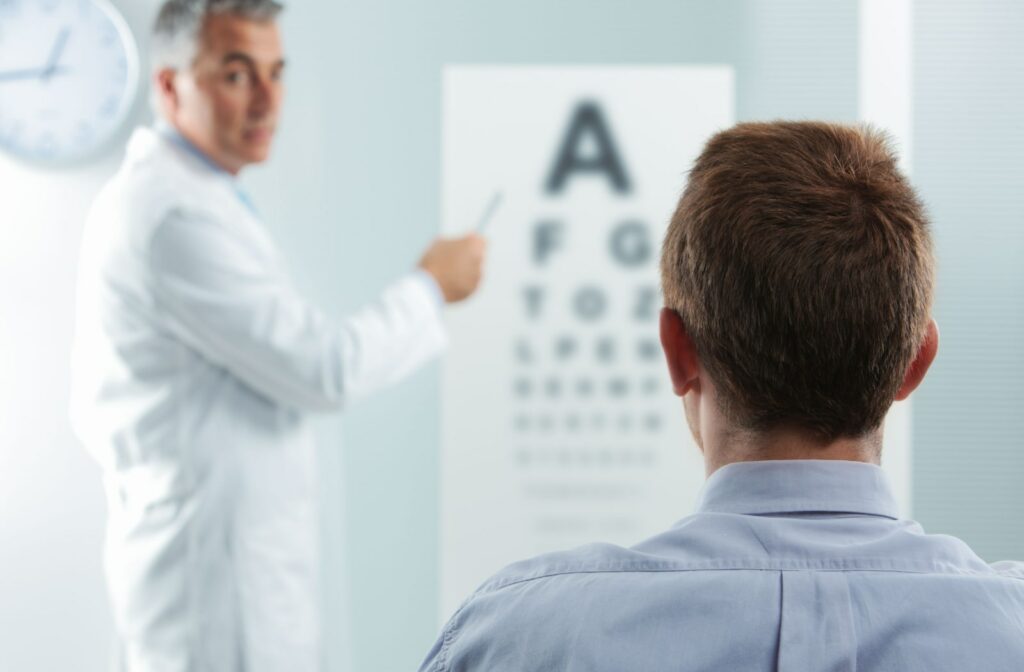 A male patient looks at a Snellen chart while his optometrist stands in front of him, pointing to the letters on the chart. The view is from the back of the patient's head.
