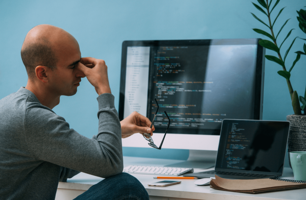 A bald man pinching the bridge of his nose and resting his eyes in front of a laptop and a computer monitor.