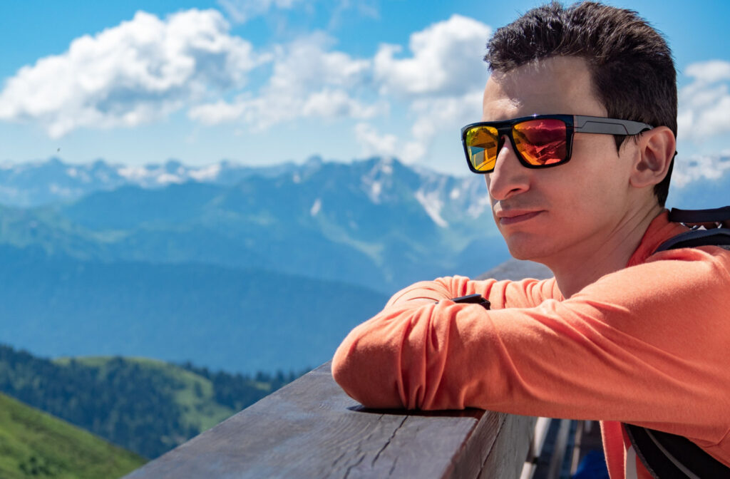A young man is wearing polarized sunglasses on a sunny day outdoors with a mountains background.