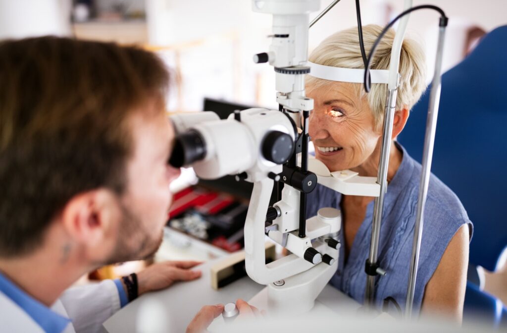 An older woman smiles as she undergoes a slit lamp exam while her optometrist inspects her eyes.