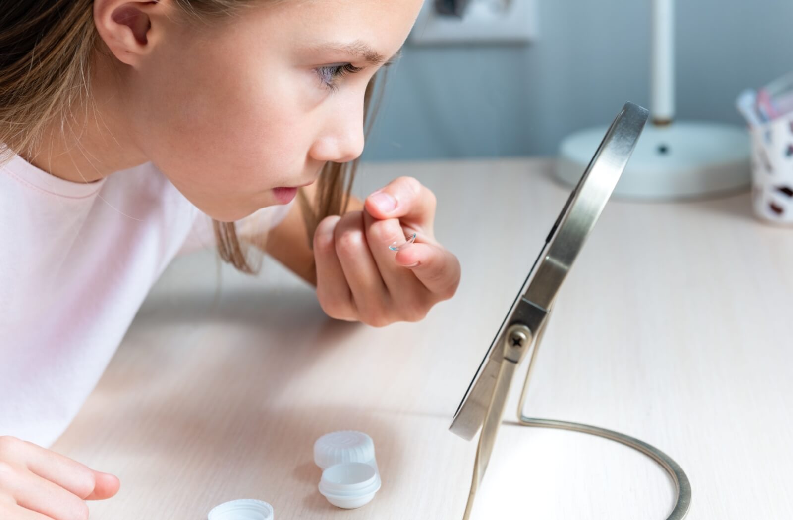 A girl holding a contact lens on her left index finger and looking in the mirror before inserting it in her eye.