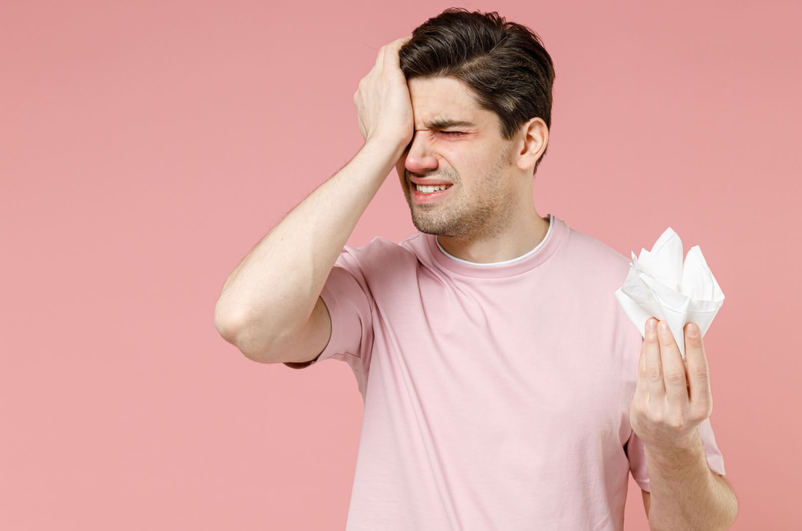 Man holding tissues and pressing his forehead, appearing uncomfortable with eye irritation.