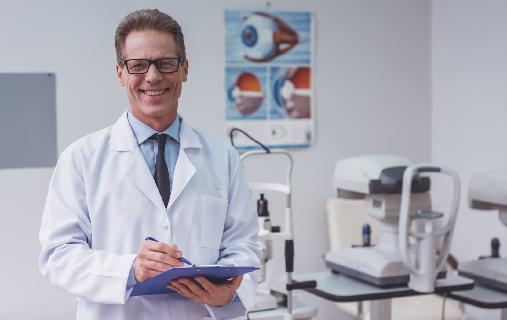 An Optometrist stands smiling in their brightly lit office, several pieces of eye exam equipment are behind them.