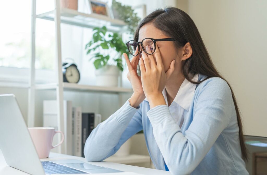 A person working at a laptop lifts their glasses to rub their irritated eyes.