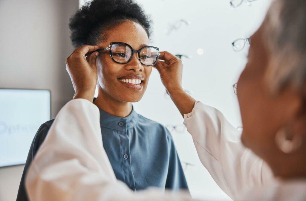 An optician helps a smiling person try on a pair of eyeglasses in an eyewear shop, adjusting the frames while display racks of glasses are visible in the background.
