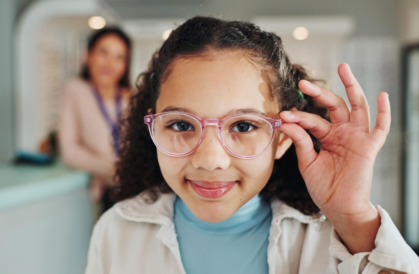 A child wearing light pink eyeglasses smiles at the camera and adjusts the frames with one hand, while an adult stands slightly blurred in the background inside an optical clinic.