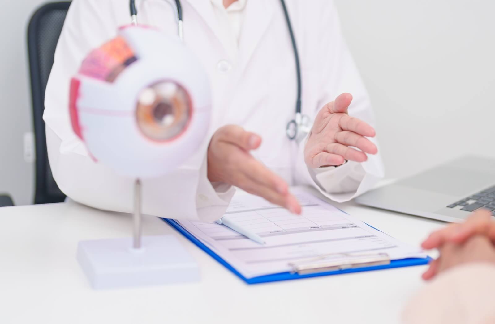 A clinician in a white coat gestures while discussing information with a patient, with an anatomical eye model and a clipboard on the desk between them.