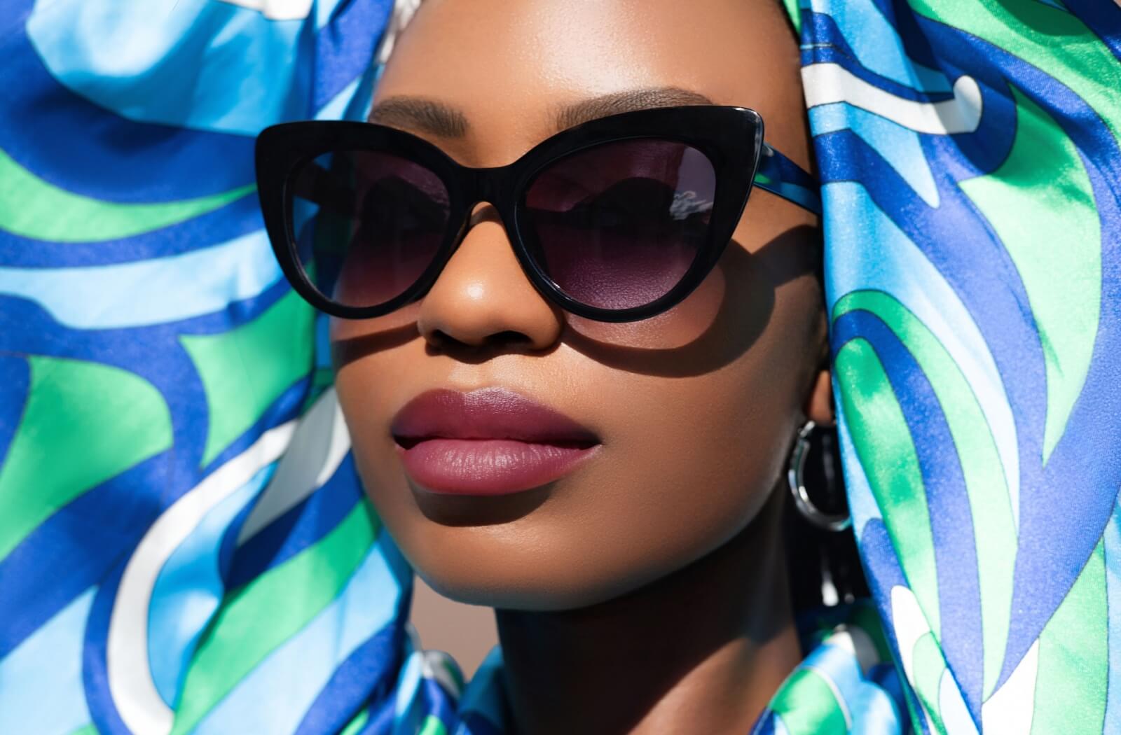 A person wearing a vibrant blue and green patterned headscarf and dark, oversized cat-eye sunglasses, showcasing 2026 feminine eyewear fashion trends.