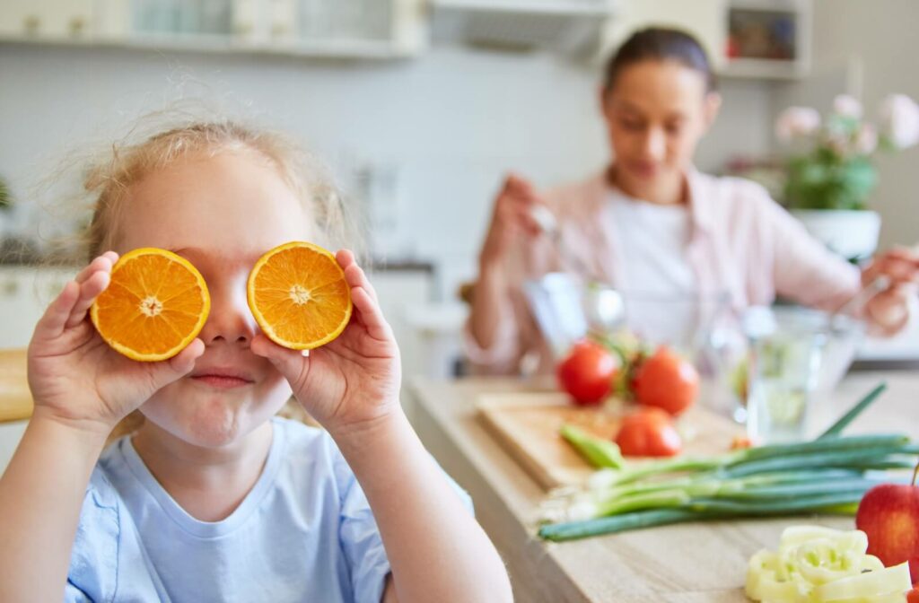 A child holding two orange slices over their eyes in a kitchen while a person prepares a healthy meal in the background, illustrating the importance of nutrition for eye health.