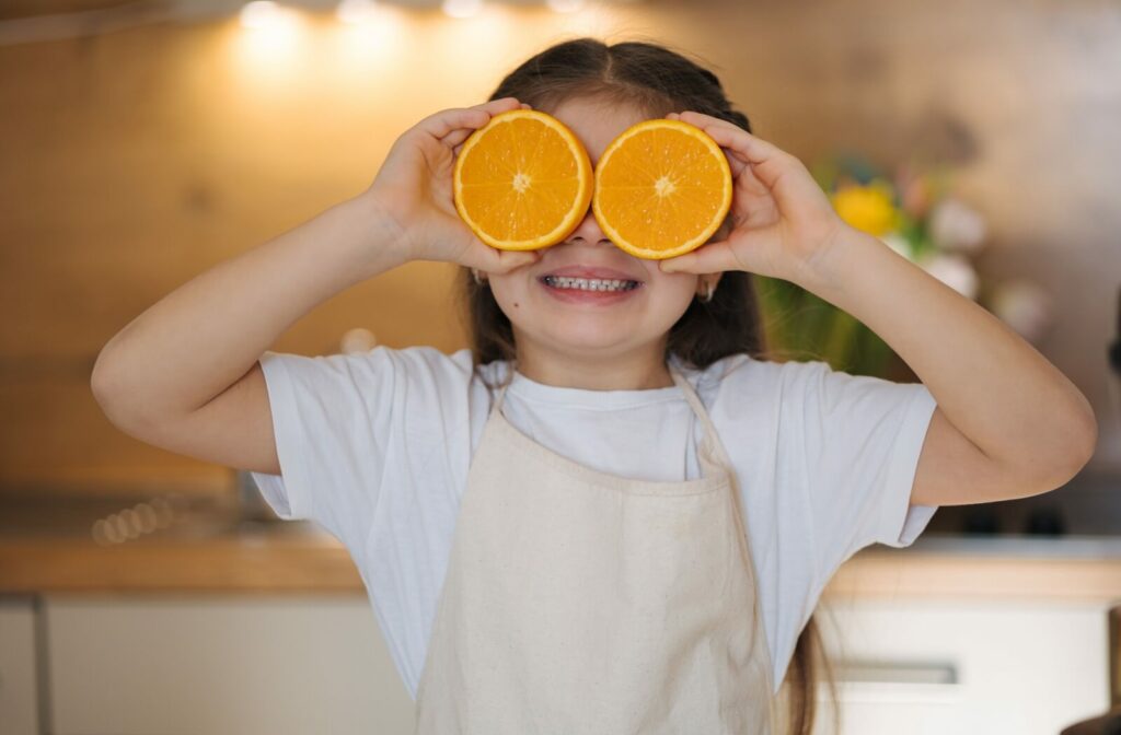 A child in a white shirt and tan apron smiling while holding two orange slices over their eyes in a kitchen, illustrating the connection between healthy nutrition and eye health.