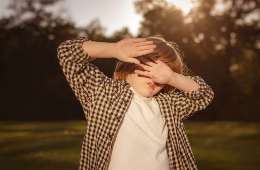 A child outdoors shielding their eyes with their hands from bright sunlight.