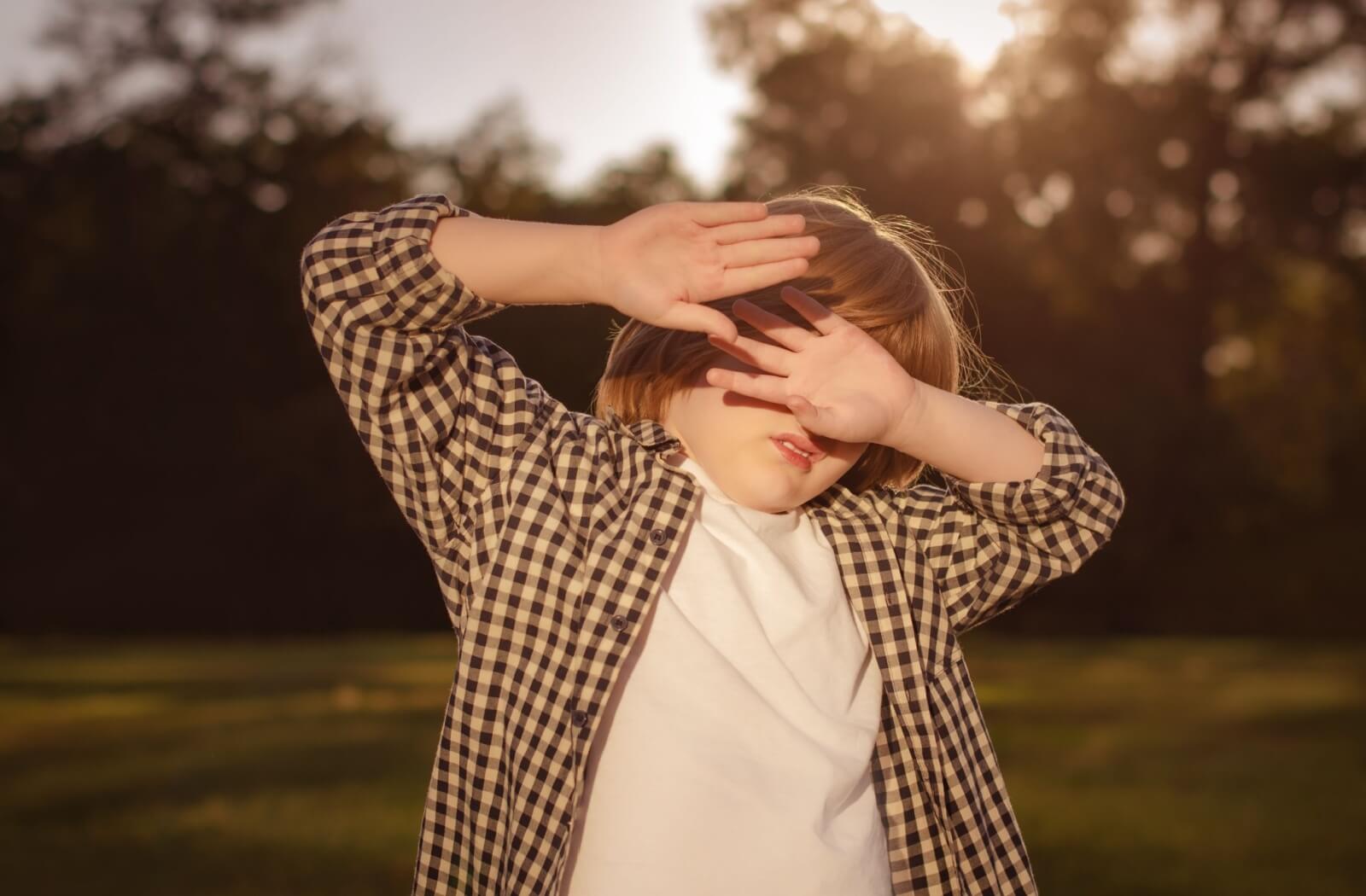 A child outdoors shielding their eyes with their hands from bright sunlight.